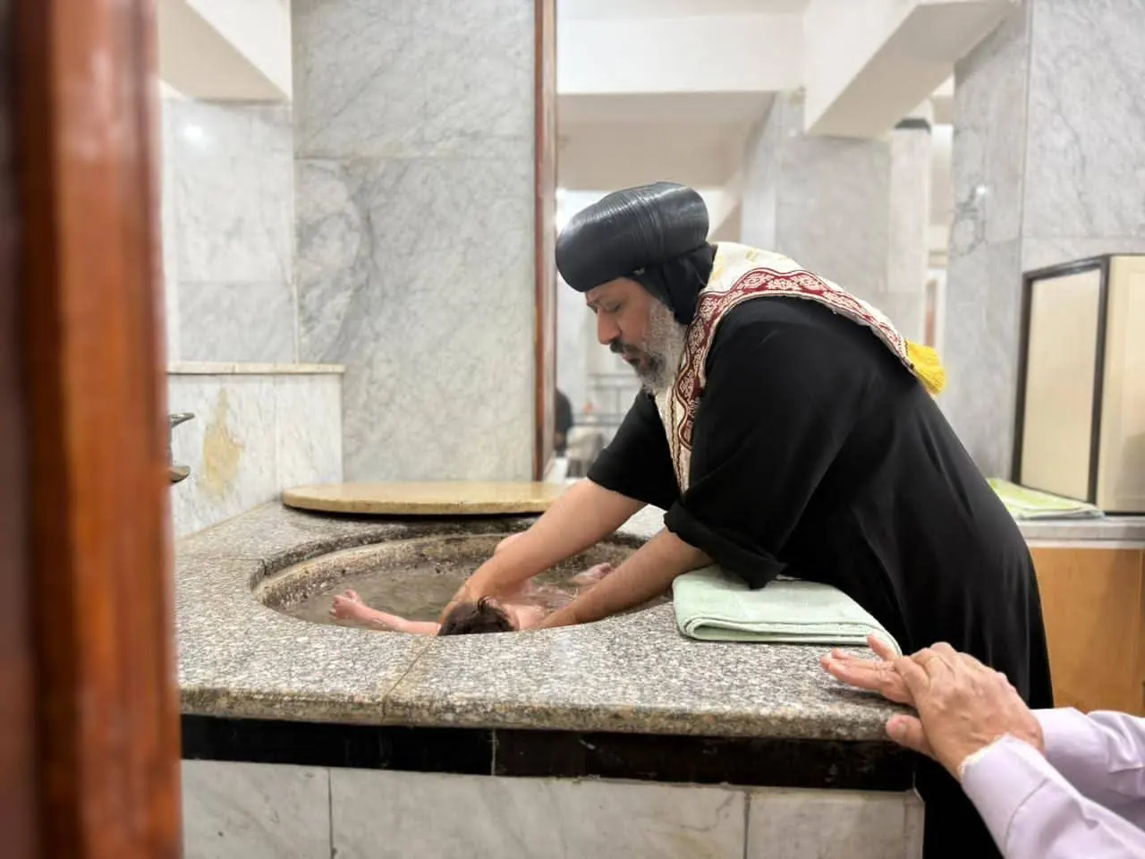 A priest in a black vestment leaning over a stone font, one hand steadying the child being baptised in it.