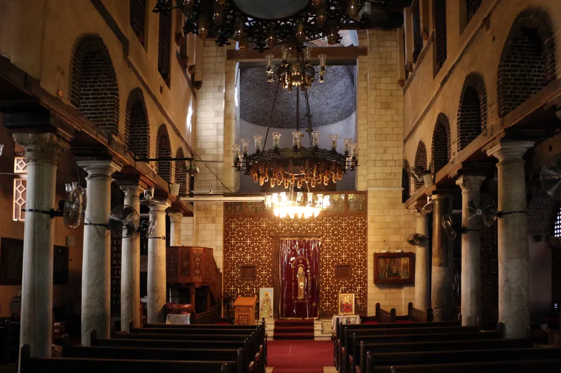The inside of an ancient church: two rows of stone columns, a dark wooden screen inlaid with crosses across the far end, and oil lamps hanging from a great chandelier.