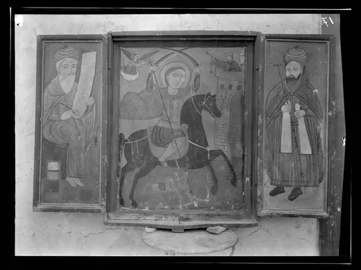 A black and white photograph of a painted wooden triptych hanging on a plastered wall, a mounted saint in the centre panel and a robed elder on each wing.