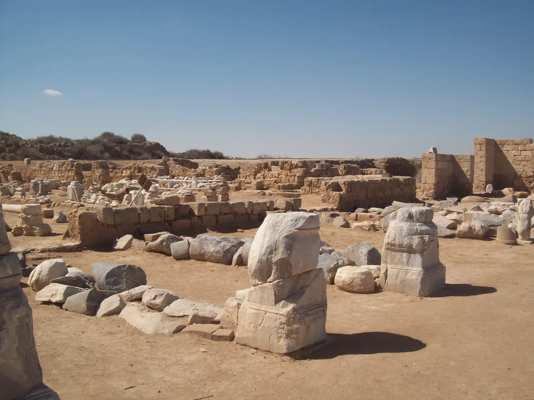Low stone walls, broken column bases and scattered blocks of white marble on flat desert sand under a clear sky.