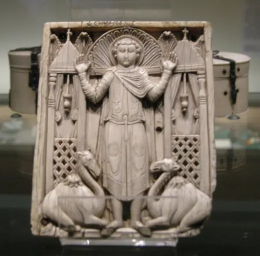 A carved ivory plaque of a soldier standing under an arch with both hands raised, a kneeling camel on each side of him.
