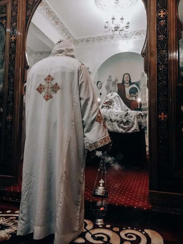 A priest in a white cope seen from behind, swinging a censer, smoke rising toward the altar.