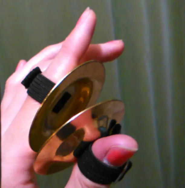 A hand holding a pair of small brass cymbals joined by elastic loops.