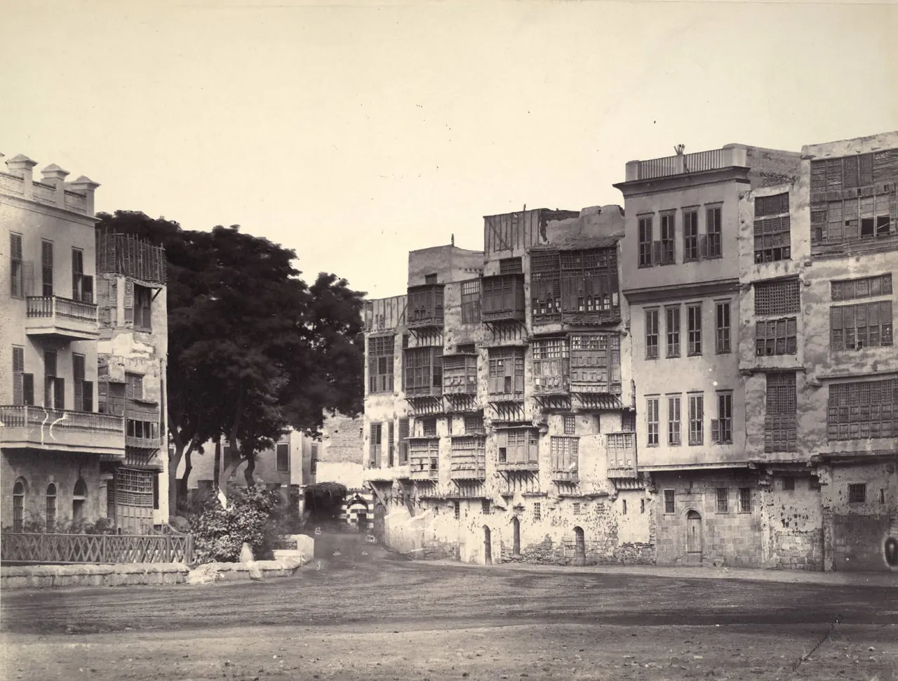 An albumen photograph of a Cairo street: tall stone houses with tiers of latticed wooden bay windows overhanging an empty dirt square.