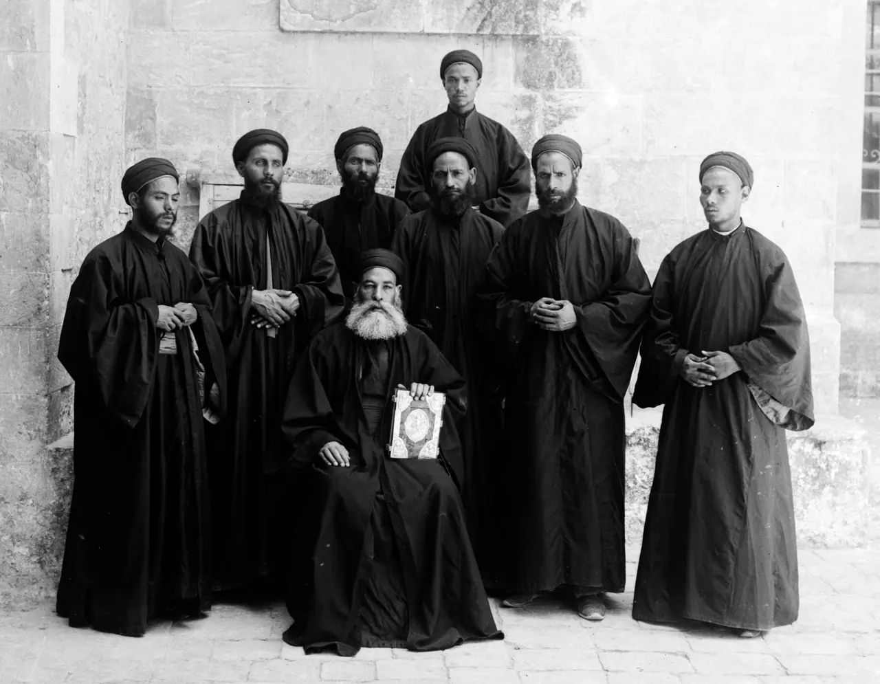 A black-and-white photograph of eight Coptic monks in black robes and turbans, one seated at the centre holding an icon, the rest standing around him against a stone wall.