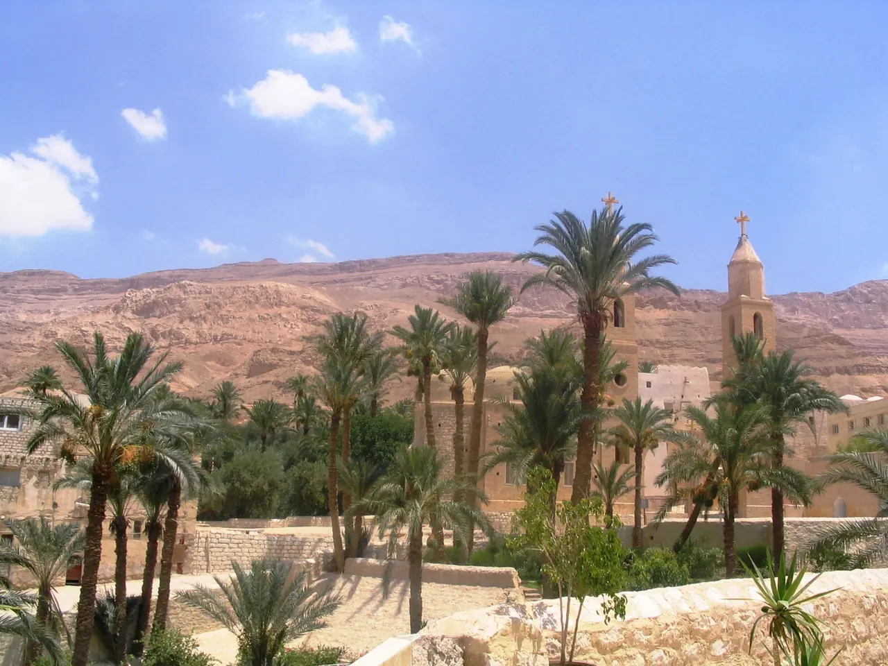 A walled monastery of pale stone with a bell tower and cross, set among date palms below a long red desert cliff.