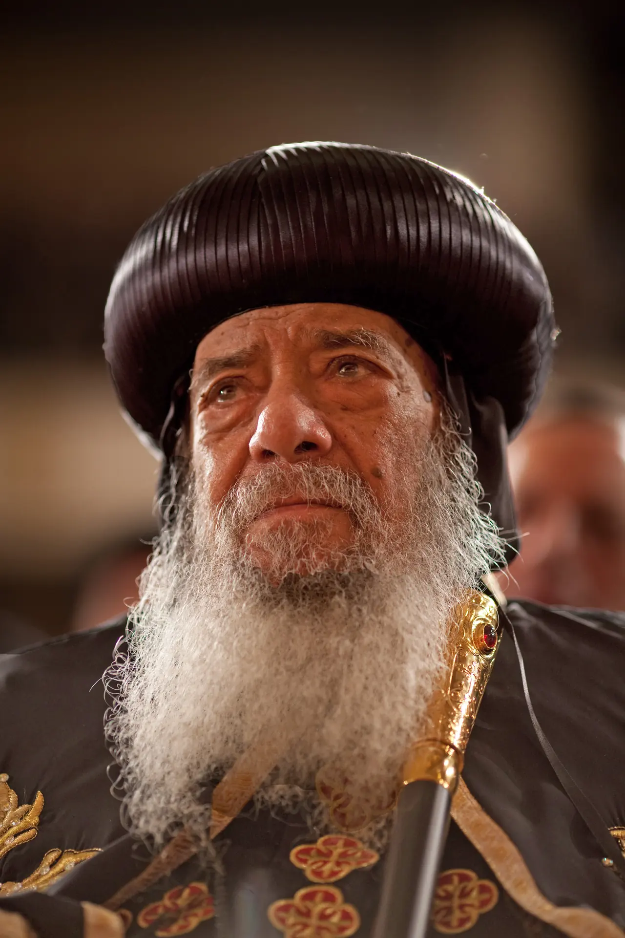 A close portrait of Pope Shenouda III, white-bearded, in a domed black headdress and gold-embroidered vestment, looking past the camera.