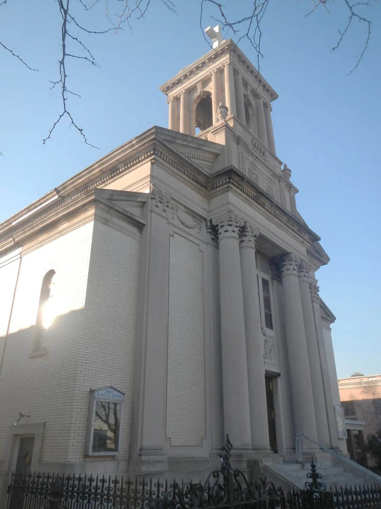A grey neoclassical church with fluted columns and a bell tower, a Coptic cross standing on top of it, seen from the street corner against a clear sky.